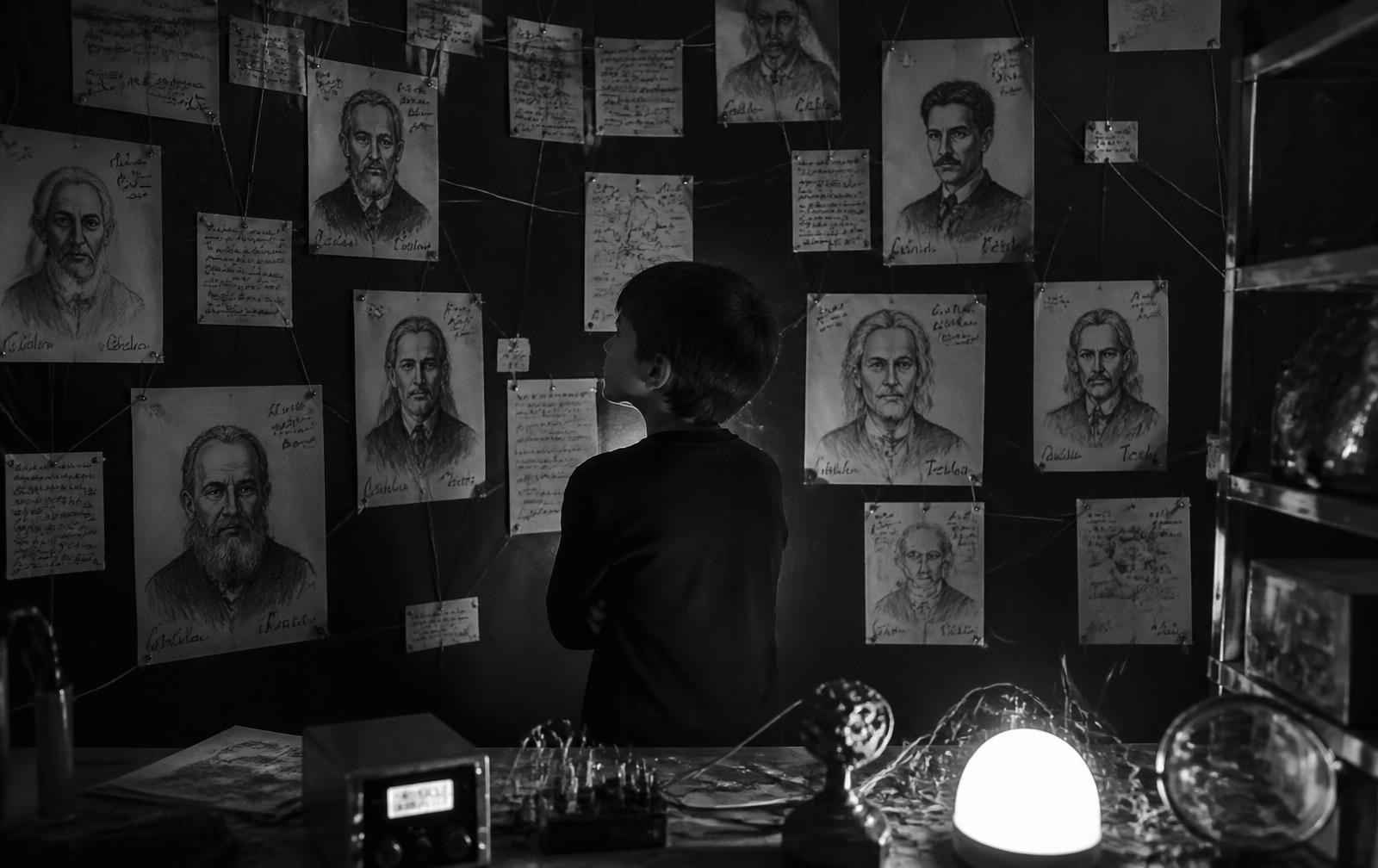 Ein Kind steht vor einer Wand aus gezeichneten Porträts, Notizen und verbundenen Fäden neben einer improvisierten Versuchsanordnung